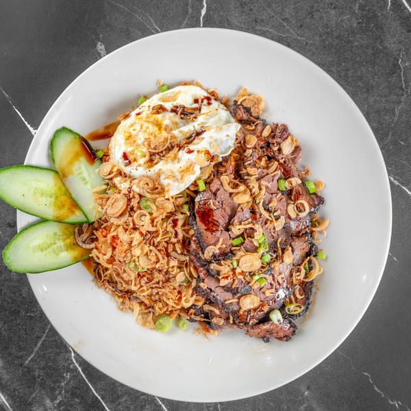 White plate with sliced beef, fried egg, crispy shallots, and cucumber slices on dark marble.