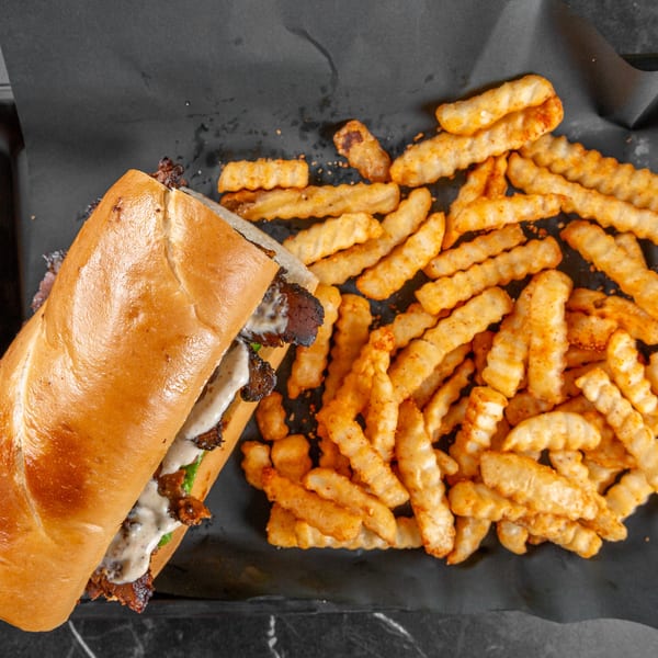 A long sub sandwich with meat and sauce next to seasoned crinkle-cut fries on a black tray.
