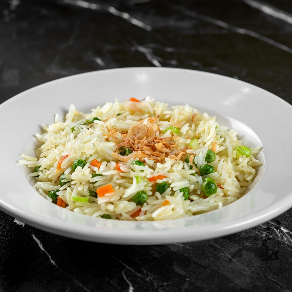 White rice with peas, carrots, and fried onions in a white bowl on a dark marble surface.